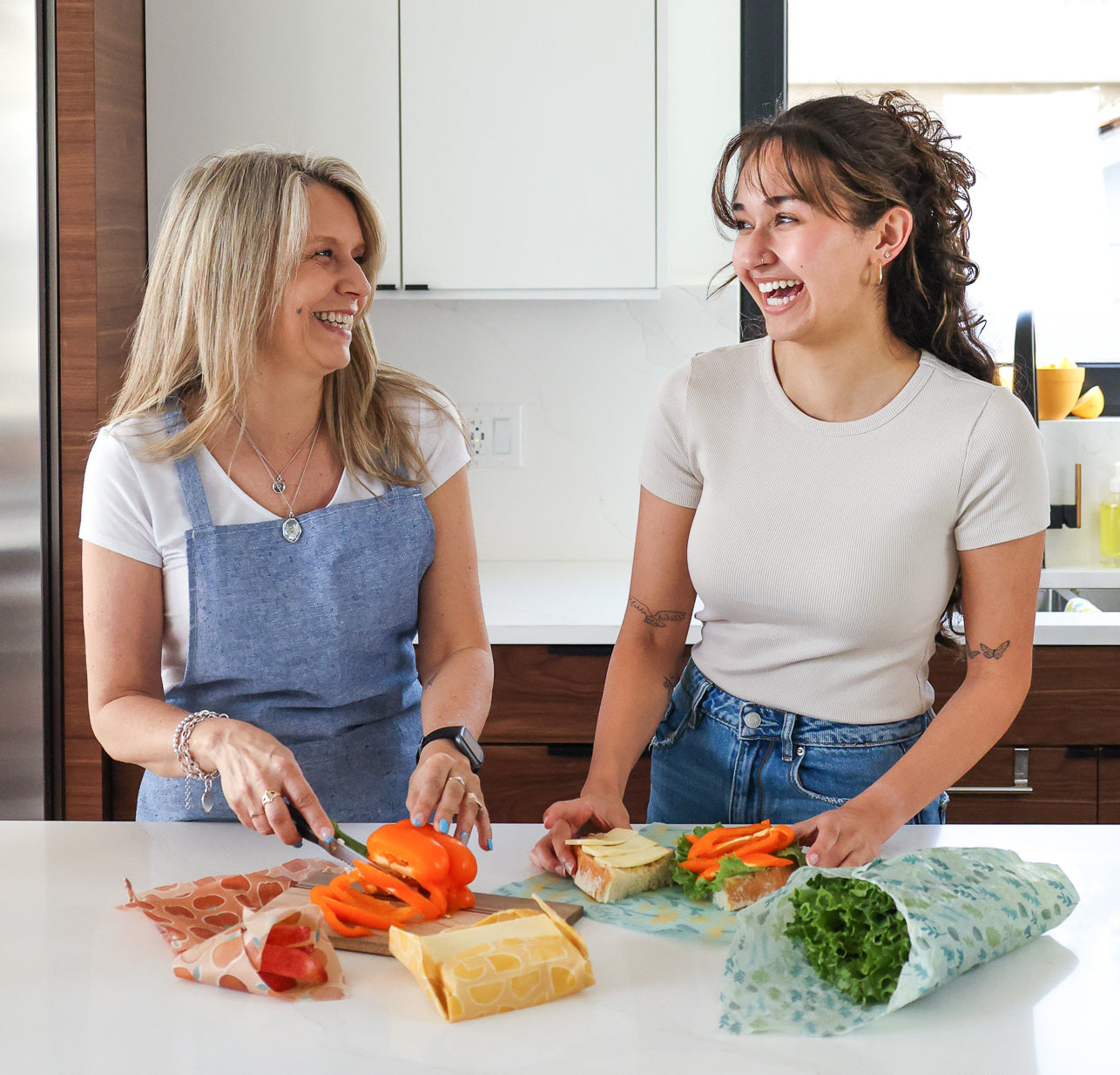 Mother and daughter in a bright kitchen preparing a sandwich with various items wrapped in nature bee beeswax wraps. This sustainable alternative to plastic wrap keeps for fresh for longer like peppers, cheese, lettuce, and is perfect for taking food like sandwiches on the go.