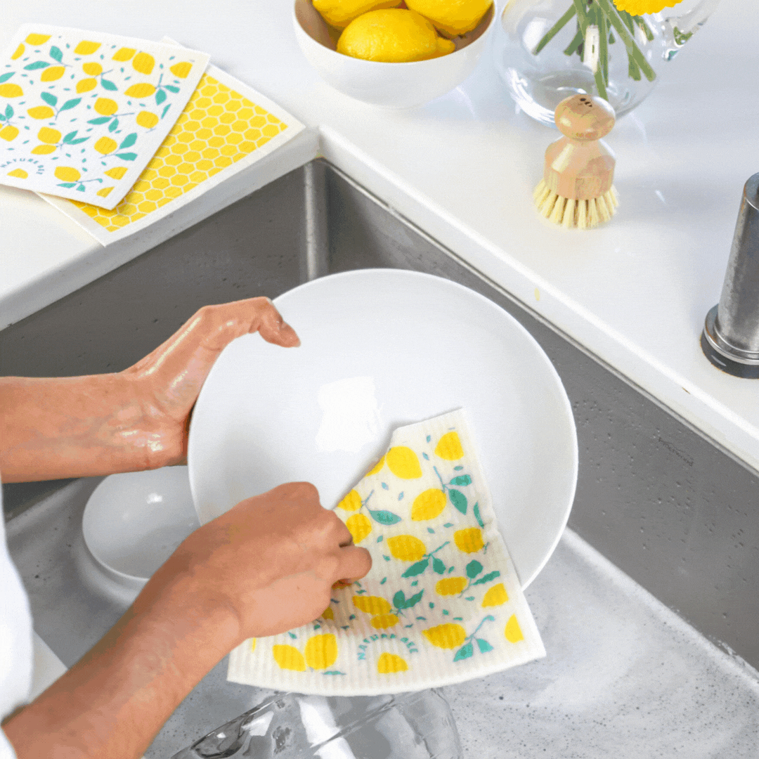 Three images rotating through a slideshow. Image 1 shows a large green bowl being washed with a swedish dishcloth in a sudsy sink. Image 2 shows spilled orange juice being wiped up by a cloth and image 3 shows cleaning a bathroom counter with a cloth and cleaning spray