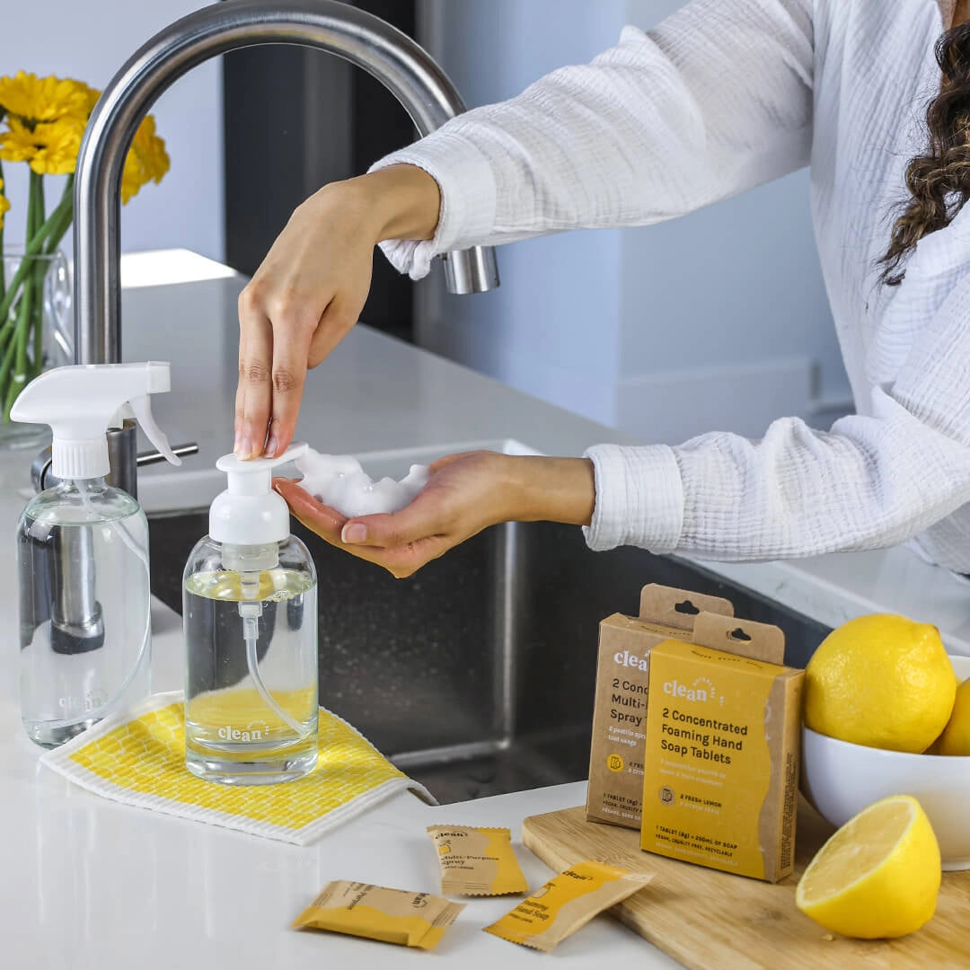 A beautiful glass foaming hand soap dispenser is being filled up with warm water from the sink, and then a foaming hand soap tablet is being dropped in and dissolves into a beautiful foamy soap. 